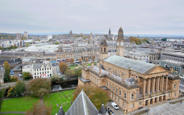 Aerial view of a historic city with a large, ornate stone building featuring columns and a clock tower in the foreground, surrounded by other buildings, trees, and a cloudy sky.