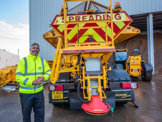 A man in a high-visibility jacket stands smiling next to a large yellow spreading vehicle with bold red and yellow warning stripes and the word "SPREADING" on the back, in an industrial yard.