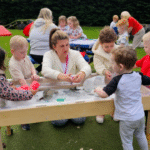 Several young children and an adult gather around an outdoor sensory table, playing with white, sand-like material. More children and adults are visible in the background, participating in similar outdoor activities.