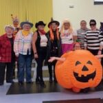 A group of people in creative Halloween costumes, including a pumpkin, witches, a clown, and other festive outfits, stand smiling together indoors.