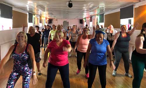 A group of women of various ages exercise together in a brightly lit studio, smiling and moving energetically in coordinated workout attire. The wooden floor reflects the light from the large windows.