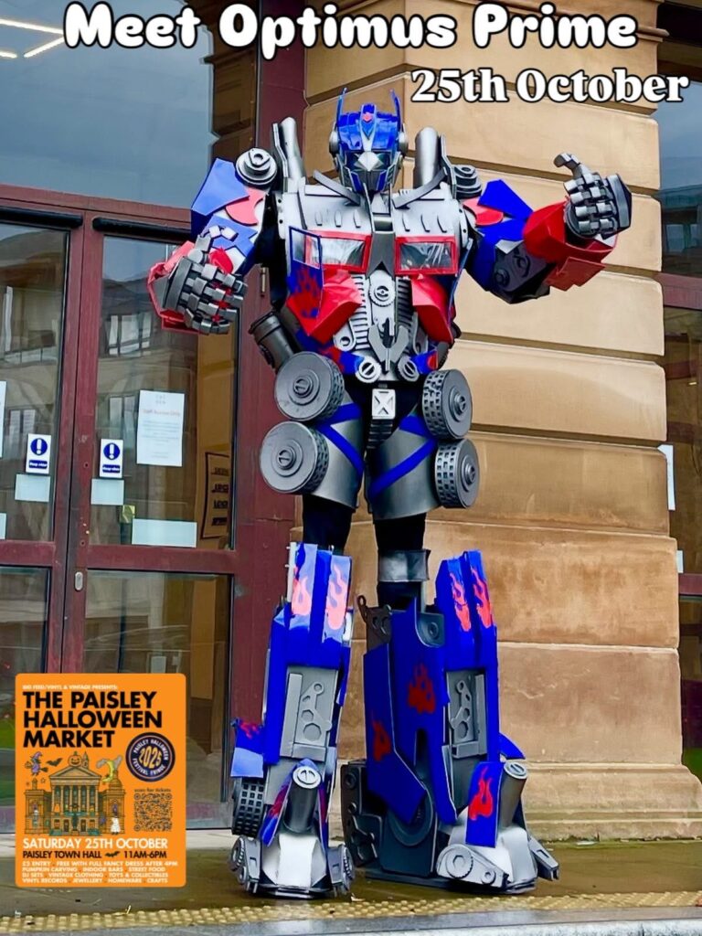 A person in an Optimus Prime costume stands outside a building. Text above reads "Meet Optimus Prime 25th October." A poster at the bottom left advertises "The Paisley Halloween Market" on Saturday 28th October.