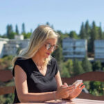 A woman with long blonde hair wearing sunglasses and a black top sits at an outdoor table, looking at her phone, with trees and buildings in the background on a sunny day.