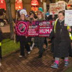 A group of people march at night holding signs and banners about women’s rights and safety, including “Not because she’s your sister, mother, daughter, but because she’s human.” Some wear high-visibility vests and winter clothing.