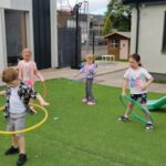 Five children play outside on artificial grass; four spin hula hoops while one girl stands on top of a playground slide. A small playhouse and modern buildings are in the background.