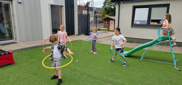 Five children play outside on artificial grass; four spin hula hoops while one girl stands on top of a playground slide. A small playhouse and modern buildings are in the background.