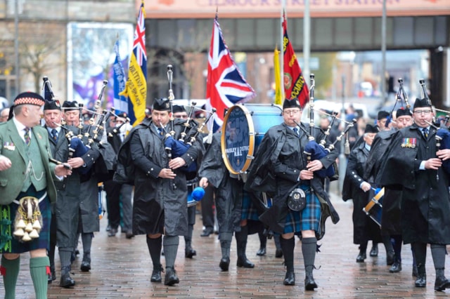 A marching band in Scottish attire plays bagpipes and drums while parading down a wet street, carrying flags. A sign reading "GLASGOW CENTRAL STATION" is visible in the background.