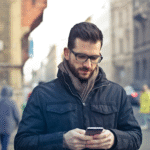 A man wearing glasses and a dark jacket stands outdoors on a city street, looking down and using his smartphone. The background shows blurred people walking and old buildings.