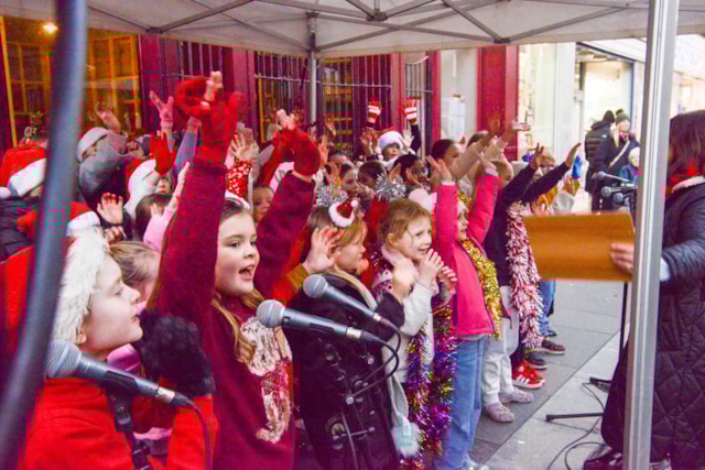 A group of children dressed in festive clothing and accessories sing together under a canopy, raising their hands enthusiastically. Microphones are set up in front, and some adults are present at the side.