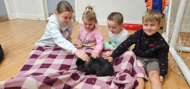 Four young children sit on a checkered blanket indoors, smiling and gently petting two small black rabbits in front of them.