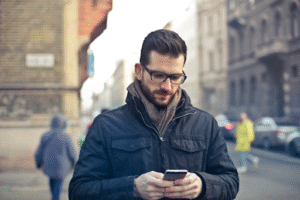 A man wearing glasses and a dark jacket stands outdoors on a city street, looking down and using his smartphone. The background shows blurred people walking and old buildings.