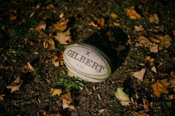 A Gilbert rugby ball lies on the ground among autumn leaves and grass, partially in shadow.