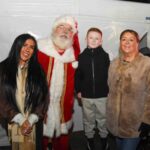 Three adults and one child pose for a photo. One adult is dressed as Santa Claus. The group stands in front of a white tent, smiling at the camera.