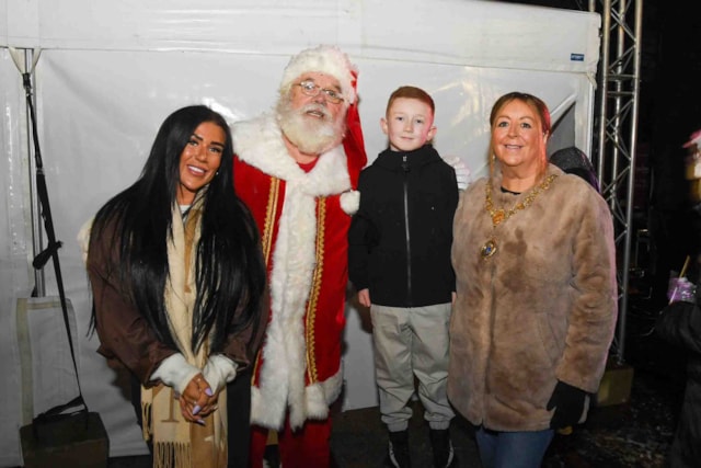 Three adults and one child pose for a photo. One adult is dressed as Santa Claus. The group stands in front of a white tent, smiling at the camera.