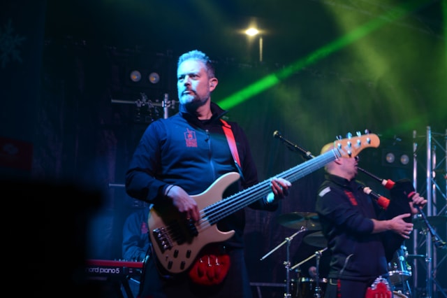 A musician playing an electric bass guitar on stage with colorful lights and another band member playing bagpipes in the background. Both are wearing dark outfits and the stage is dimly lit.