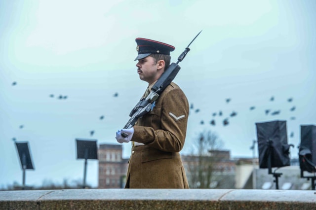 A uniformed guard with a rifle stands at attention outdoors, wearing a peaked hat and white gloves. Birds are flying in the cloudy sky behind him, with buildings and blurred monuments in the background.