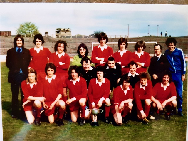 A vintage photo of a boys’ soccer team in red uniforms posing on a grassy field with two adults in tracksuits. A trophy sits in front of them. Stands and a building appear in the background.