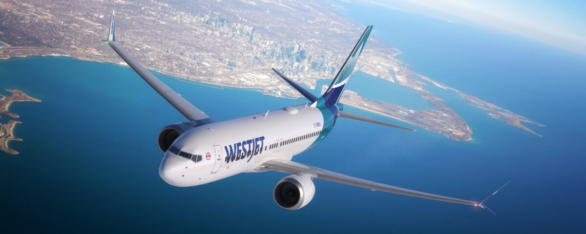A WestJet airplane flies over a large body of water and land, with a city skyline visible in the background under a clear, blue sky.