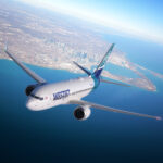 A WestJet airplane flies over a large body of water and land, with a city skyline visible in the background under a clear, blue sky.