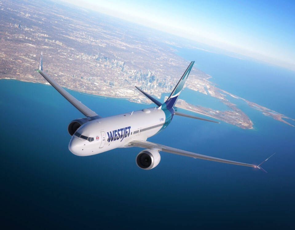 A WestJet airplane flies over a large body of water and land, with a city skyline visible in the background under a clear, blue sky.