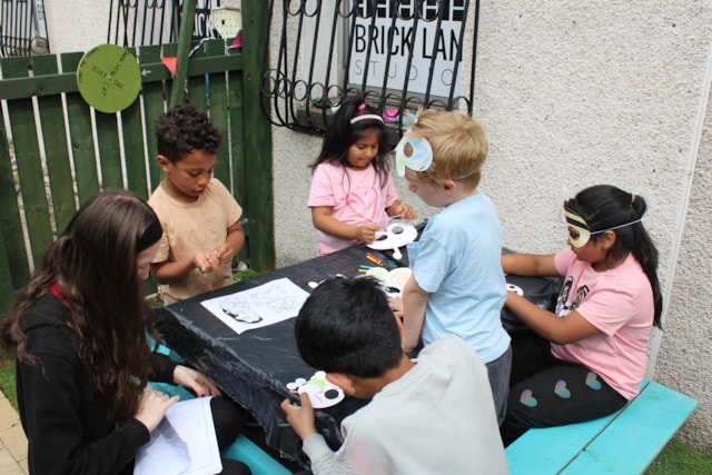 A group of young children and an adult sit and stand around a table outdoors, engaged in arts and crafts activities. Some of the children are wearing animal masks and coloring or assembling paper crafts.