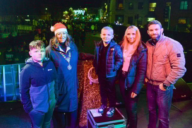 A group of five people, including a woman in a festive hat and a mayoral chain, pose on a stage at night with colorful lights and a crowd in the background.