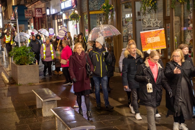 A group of people walk down a wet city street at night, some holding transparent umbrellas. Many wear coats and a few have reflective vests. Shop signs and lights line the street, and signs reading "FREEDOM" are visible.