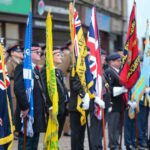 A group of people in military uniforms stand in a row holding colorful flags and banners, including the Union Jack, at an outdoor ceremony on a city street. Others in uniform are visible in the background.