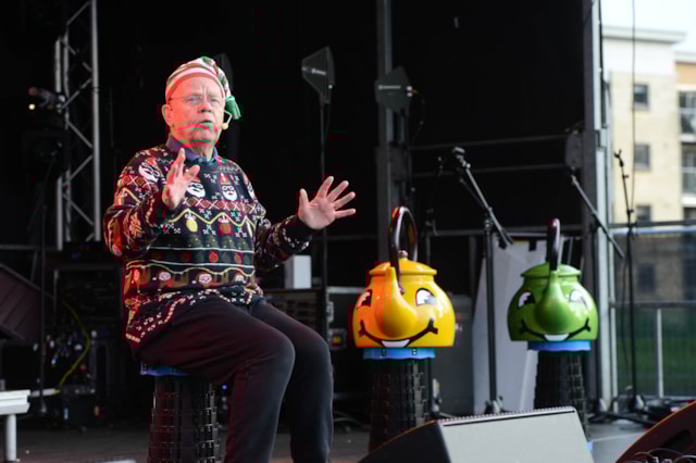 An elderly man in a festive sweater and striped elf hat sits on stage, gesturing with his hands. Two large, colorful buckets with smiling faces are beside him. Stage equipment and buildings are visible in the background.