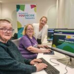 Three adults sit at computers in an office, smiling at the camera. A colorful banner behind them reads "Renfrewshire YOUDECIDE Tenants." One woman stands, while two others sit and use desktop computers.