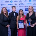 Four people dressed formally stand together at an award event. Two women in dresses hold a framed certificate and a trophy. A man in a tuxedo and another woman smile beside them. Event sponsor logos are displayed on the backdrop.