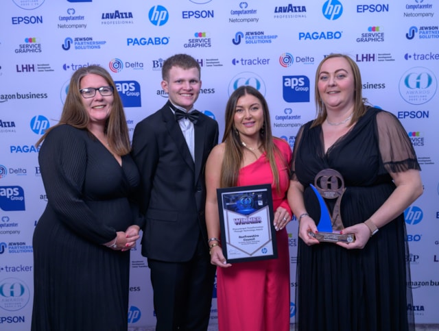 Four people dressed formally stand together at an award event. Two women in dresses hold a framed certificate and a trophy. A man in a tuxedo and another woman smile beside them. Event sponsor logos are displayed on the backdrop.