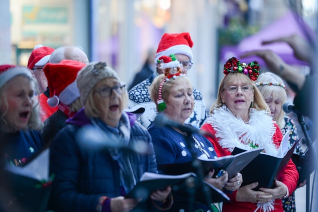 A group of people wearing festive hats and holiday accessories sing from songbooks, likely performing Christmas carols outdoors. Microphones and blurred decorations are visible in the foreground.