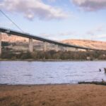 A large bridge with tall pillars spans a wide river, with hills and scattered trees in the background under a partly cloudy sky. There is sandy ground and old wooden posts in the foreground by the water’s edge.
