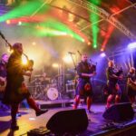 A band wearing kilts performs on an outdoor stage with colorful lights. Members play bagpipes, drums, and guitar, while fog and vibrant lighting create a lively concert atmosphere.