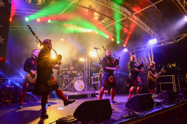 A band wearing kilts performs on an outdoor stage with colorful lights. Members play bagpipes, drums, and guitar, while fog and vibrant lighting create a lively concert atmosphere.