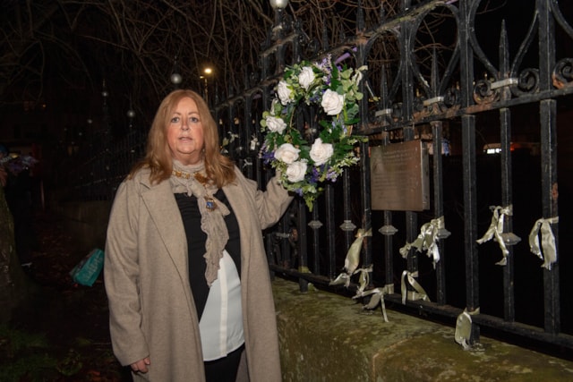 A woman stands by a metal fence at night, holding a floral wreath. Ribbons are tied to the fence, and a plaque is visible behind her. She wears a beige coat and white blouse. Trees with bare branches are in the background.