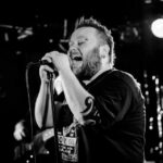 A bearded man with a visible tattoo on his arm passionately sings into a microphone on stage. The black and white photo captures him mid-performance, with stage lights and a blurred guitarist in the background.