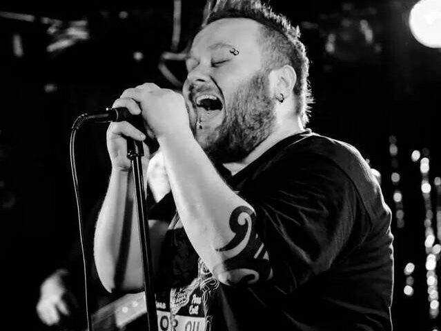 A bearded man with a visible tattoo on his arm passionately sings into a microphone on stage. The black and white photo captures him mid-performance, with stage lights and a blurred guitarist in the background.