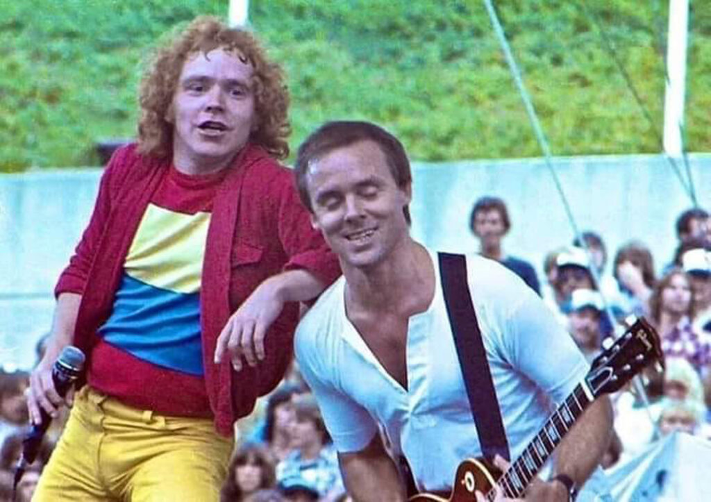 Two musicians perform on an outdoor stage; one is singing with a microphone, wearing a colorful shirt and yellow pants, while the other plays a guitar, with a crowd watching in the background.