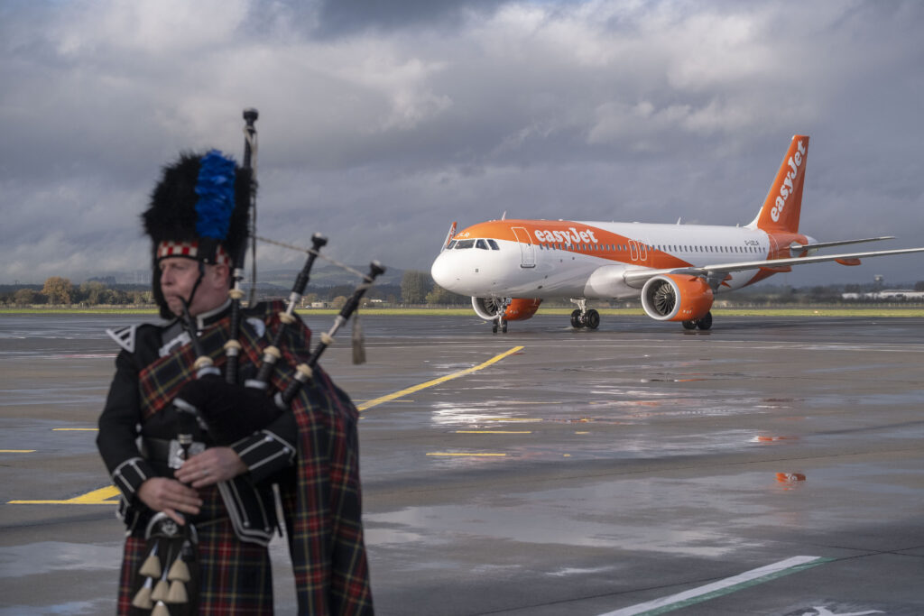 A bagpiper in traditional Scottish attire stands on a wet airport tarmac, while an orange and white easyJet airplane taxis in the background under a cloudy sky.