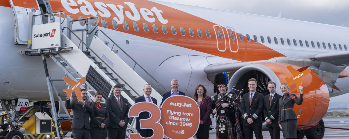 A group of easyJet crew and others stand in front of an easyJet airplane holding signs celebrating 30 years of flights from Glasgow, with one person dressed in traditional Scottish attire and playing bagpipes.