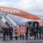 A group of easyJet crew and others stand in front of an easyJet airplane holding signs celebrating 30 years of flights from Glasgow, with one person dressed in traditional Scottish attire and playing bagpipes.