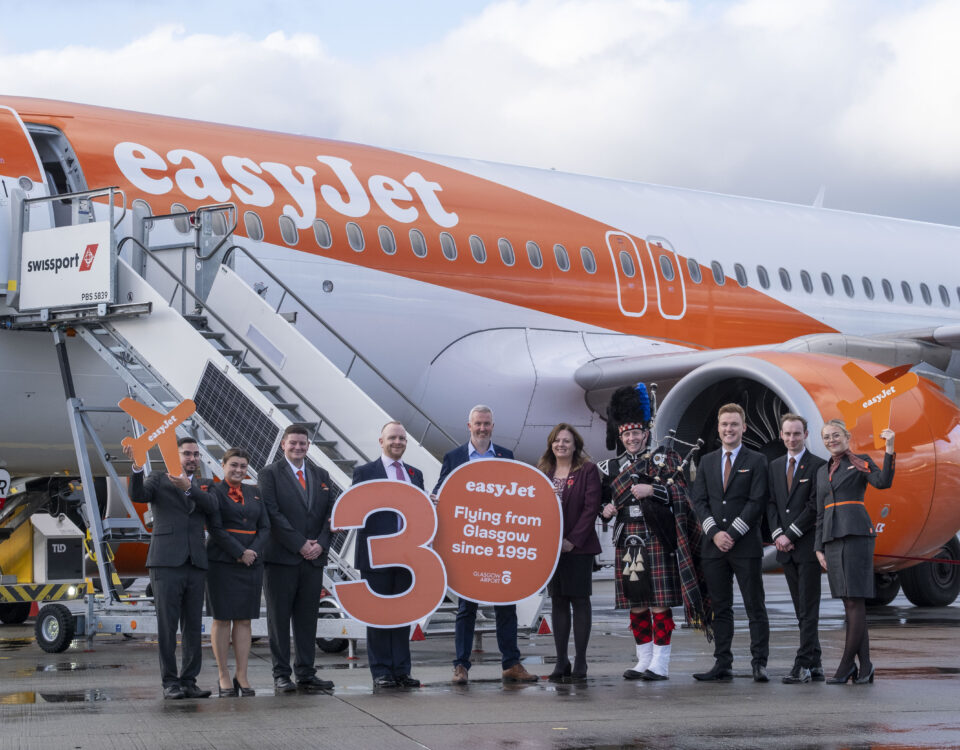 A group of easyJet crew and others stand in front of an easyJet airplane holding signs celebrating 30 years of flights from Glasgow, with one person dressed in traditional Scottish attire and playing bagpipes.