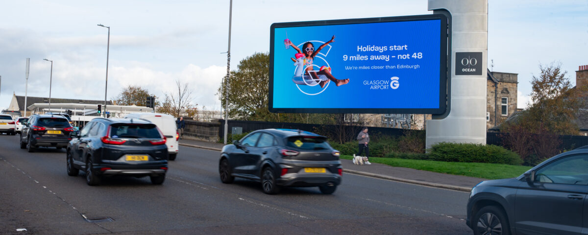 Cars driving on a road next to a large digital billboard advertising Glasgow Airport, which reads, "Holidays start 9 miles away – not 48. We’re closer than you think. Glasgow Airport.