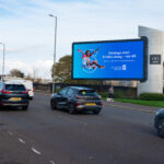 Cars driving on a road next to a large digital billboard advertising Glasgow Airport, which reads, "Holidays start 9 miles away – not 48. We’re closer than you think. Glasgow Airport.