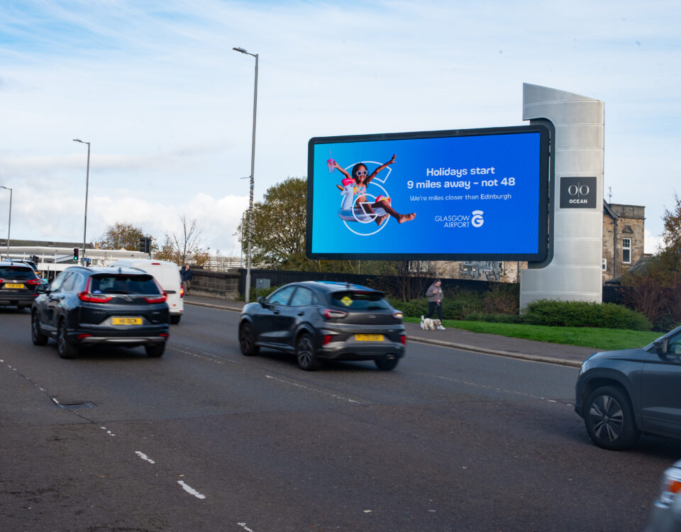 Cars driving on a road next to a large digital billboard advertising Glasgow Airport, which reads, "Holidays start 9 miles away – not 48. We’re closer than you think. Glasgow Airport.