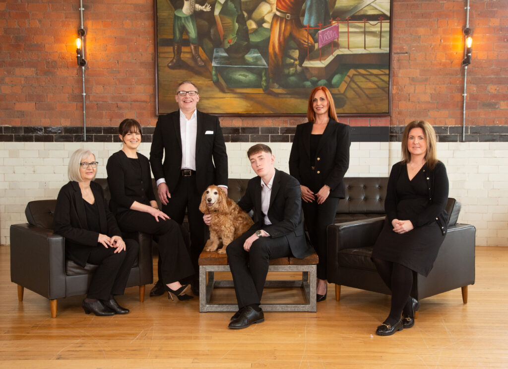 Six adults in black outfits pose indoors on dark sofas and a bench, with a brown and white dog seated at the center. They are in front of a brick wall with a large painting and two wall lights.