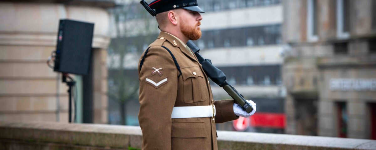 A uniformed soldier stands at attention outdoors, holding a rifle across his chest. He wears a brown dress uniform, white gloves, a white belt, and a peaked cap, with buildings visible in the blurred background.