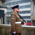 A uniformed soldier stands at attention outdoors, holding a rifle across his chest. He wears a brown dress uniform, white gloves, a white belt, and a peaked cap, with buildings visible in the blurred background.